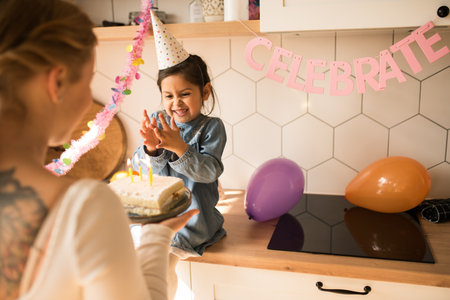 Mother holding plate with birthday cake near daughter while wishes happy birthdayの写真素材