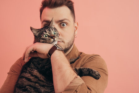 Man posing with his striped cat at the hands with pink wall at the backgroundの写真素材