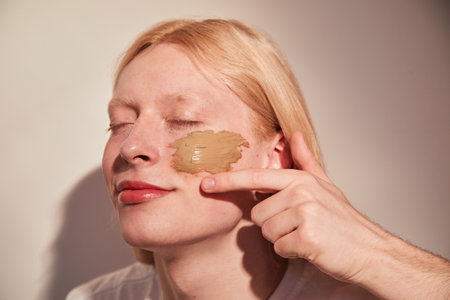 Feminine man applying concealer at his chick while posing with closed eyesの写真素材