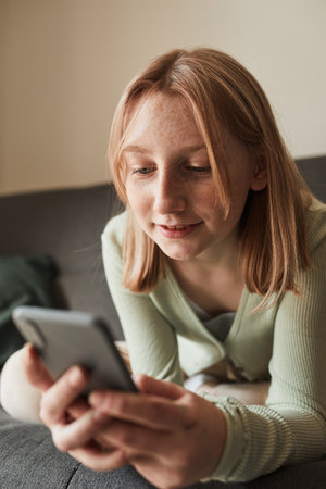 Freckled girl looking at the smartphone screen while sitting at the sofaの写真素材