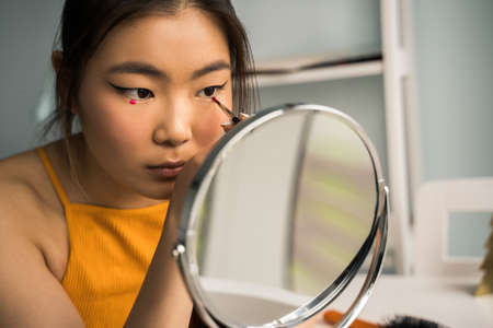 Woman with brunette hair painting her eyes and preparing creative makeup while sitting at the tableの写真素材