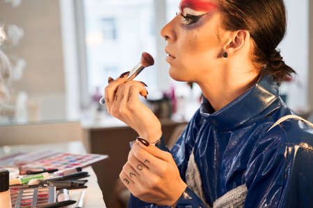 Drag queen applying blushes at his face while sitting at the dressing table
