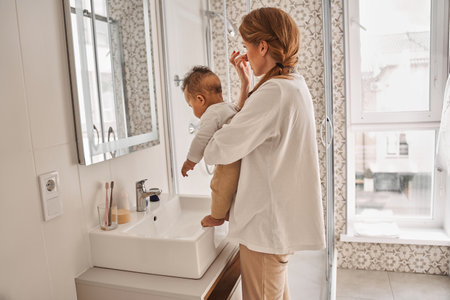Woman holding her child while applying moisturizer at her faceの写真素材