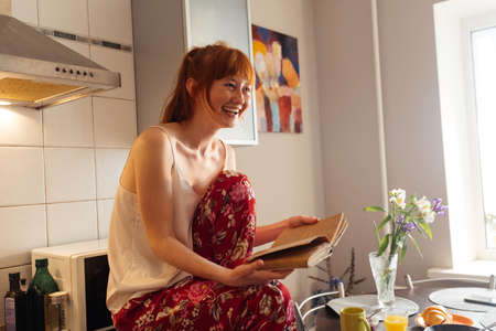 Woman sitting at comfortable kitchen, reading interesting book and laughing