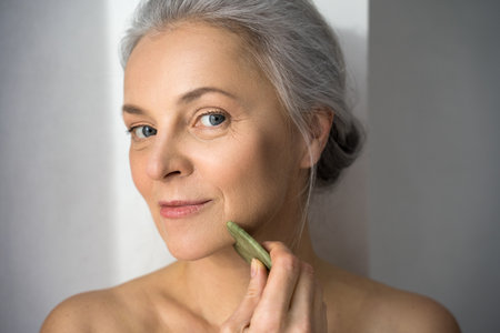 Portrait view of smiling mature grey haired woman using quartz face massager for skin treatment while looking at the camera at studio behind the white wall. Stock photoの写真素材