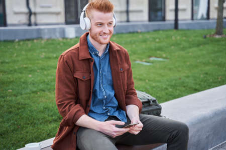 Man wearing headphones sitting on the curb and looking away with pleasure smileの写真素材