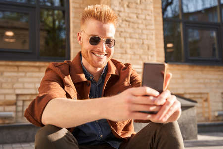 Man wearing sunglasses sitting on the pavement and looking at his smartphone