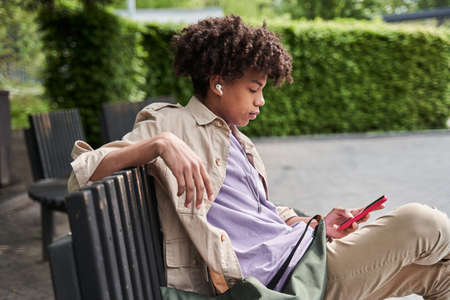 Man wearing earphones sitting at the bench at the street and listening music