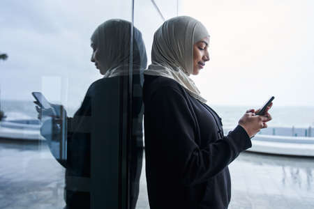 Lady in hijab holding mobile phone and looking at the screen with smile