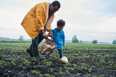 Man and his little son wearing raincoats cleaning field and picking up paper bagsの写真素材