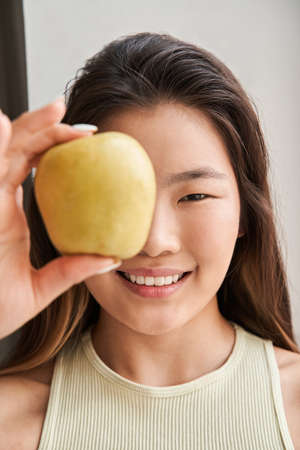 Girl smiling toothy and looking at the camera while hiding her eye behind the apple