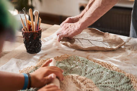 Sculptor giving a shape to the clay plate while his student holding rolling pin