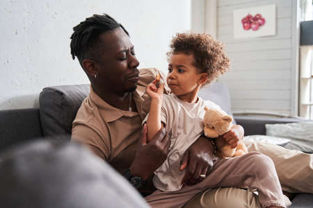 Girl playing with her father while sitting at the sofa and holding her favorite bearの写真素材