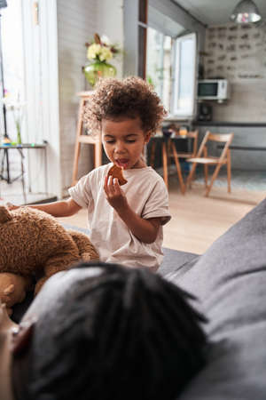 Girl eating cookie while sitting at sofa near her father and looking at him with interestの写真素材