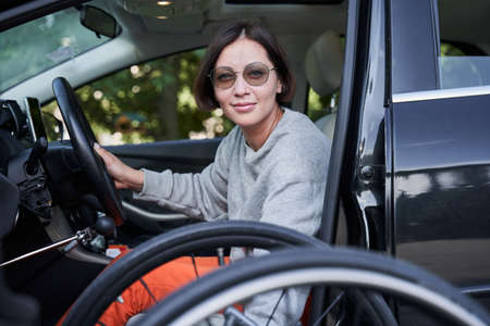 Woman wearing sunglasses folding her wheelchair into the car and smiling to the cameraの写真素材