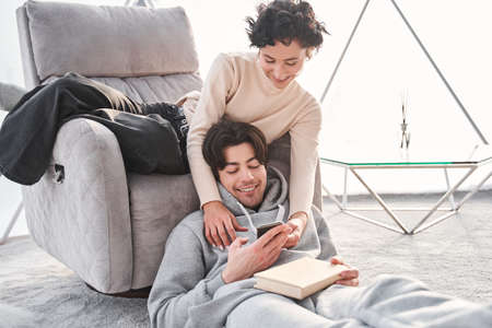 Man reading a book while his girlfriend showing to him something at smartphone