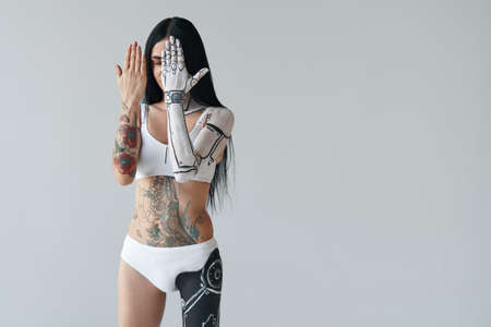 Horizontal shot of women with painted hand of robot body art. Woman posing at the studio and hiding her face behind her hands. White wall at the backgroundの写真素材