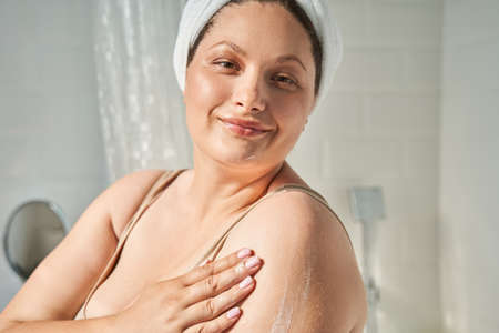 Woman looking in the camera with pleasure smile while using moisturizing cream at her bodyの写真素材