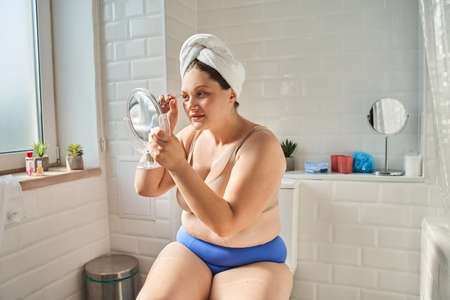 Woman is sitting at the bathroom and using tweezers while holding hand mirrorの写真素材