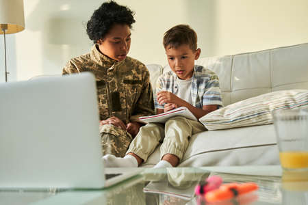 Woman wearing military jacket talking with her little son while sitting at the sofaの写真素材