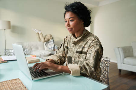 Female soldier sitting at the table and using laptop while her little son jumpingの写真素材
