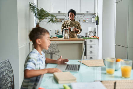 Boy sitting at the table and using laptop while his mother preparing breakfastの写真素材