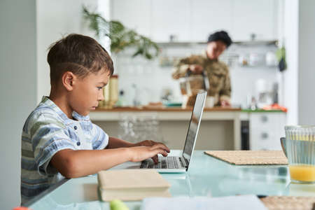 Boy typing at the laptop keyboard while his mother preparing breakfastの写真素材