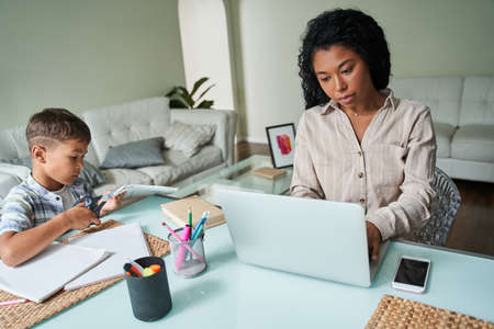 Boy drawing at the table near his multiracial mother working at the laptopの写真素材
