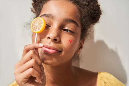 Teen girl hiding her eye behind sweet candy and looking away while posing to the camera