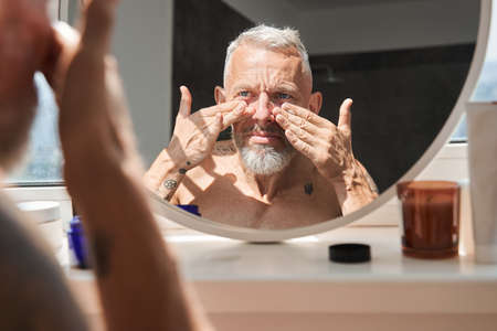 Grey haired senior man looking and examining his face at the mirrorの写真素材