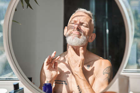 Man looking at the mirror and putting moisturizer at his neck with pleasure smileの写真素材