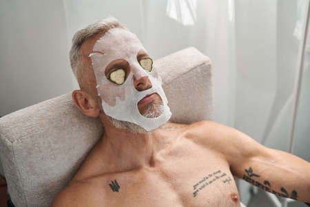 Man with cosmetic cloth mask and cucumber on faceの写真素材