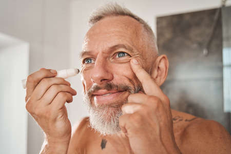 Man looking at the mirror while using moisturizing roller for skin around eyesの写真素材