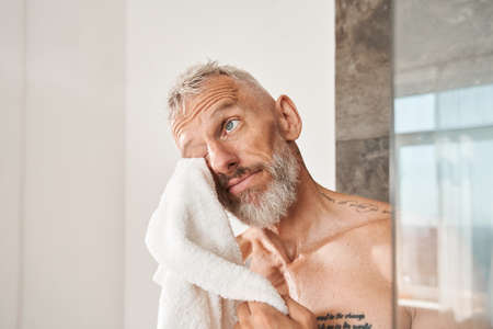 Mature bearded man wiping his face with white towel at the morningの写真素材