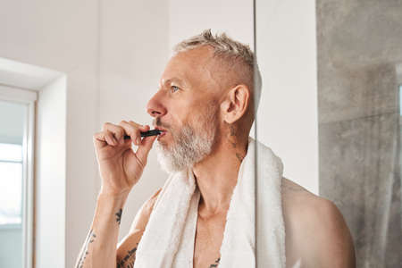 Bearded man with white towel at his neck brushing teeth at the morningの写真素材
