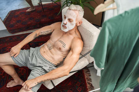 Senior man with facial mask sitting at the armchair and relaxing at homeの写真素材