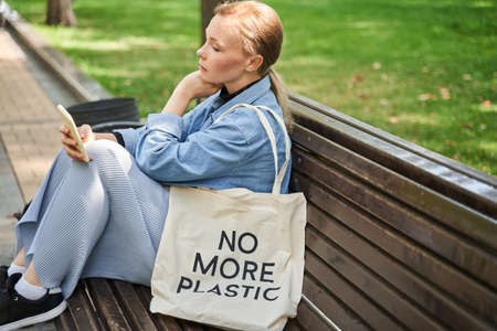 Woman sitting at the bench and using her smartphone while walking with her eco bagの写真素材