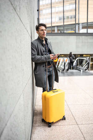 Businessman standing alone at airport while listening music and carrying his suitcaseの写真素材
