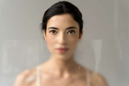 Shiny woman standing behind the transparent glass and looking straight to the camera
