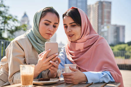 Two female person wearing headscarf spending time at their gadget while spending time