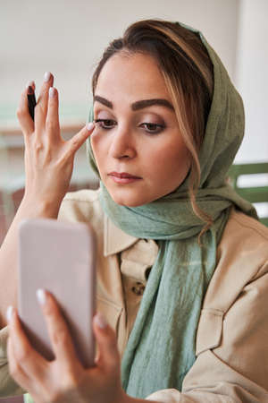 Islamic woman wearing headscarf paints her eyelashes and looking at the selfie camera at her cell