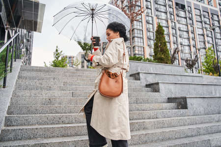 Woman holding transparent umbrella while walking outside with cup of hot coffee