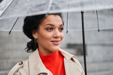 Brunette woman smiling while standing at the street with umbrella and looking away