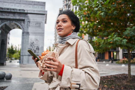 Woman looking away and eating her lunch while holding smartphone at the autumn street