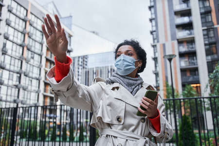 Woman wearing protective mask holding her smartphone and using app while catch a taxi.