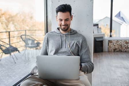 Man sitting at the windowsill at home and using his laptop with pleasure smile
