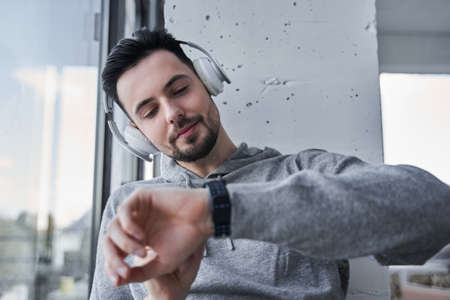 Guy looking time at his wristwatch while sitting near the window and listening musicの写真素材