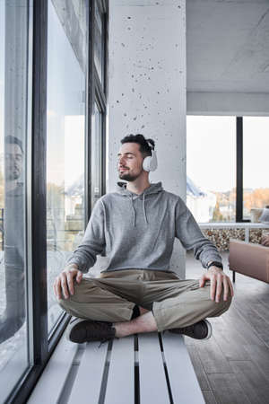 Man wearing headphones keeping eyes closed and sitting on windowsill in a lotus positionの写真素材