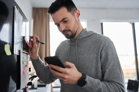 Man looking at his smartphone while writing at the colorful sticker glued to the fridgeの写真素材