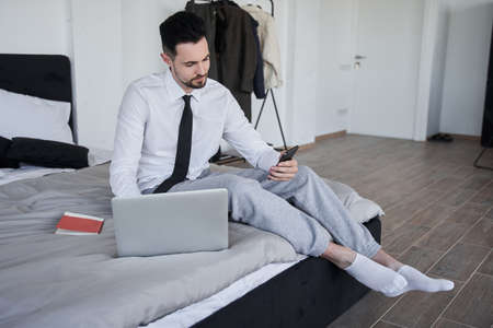 Businessman sitting at the bed and looking at the smartphone screenの写真素材